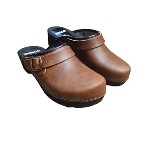 Puede incluir: Un par de zuecos de cuero marrón con cierre de hebilla y suela de madera. La palabra "BRESCO" está impresa en el lateral. Los zapatos están diseñados para la comodidad y el estilo.