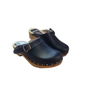 Puede incluir: Un par de zuecos de cuero negro con suelas de madera. Los zuecos tienen una correa con hebilla y están adornados con pequeños tachuelas doradas alrededor de la base. La marca "BRESCO" es visible en la plantilla.