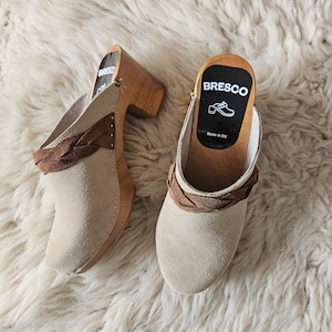 Puede incluir: Un par de zuecos de ante beige con correas de cuero marrón y tacones de madera. Los zuecos tienen una etiqueta en el talón que dice "BRESCO Made in EU".