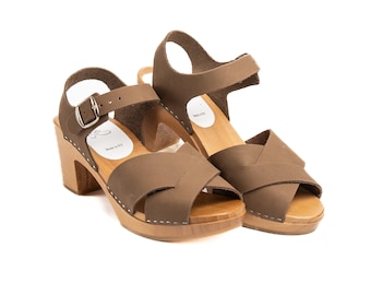 Puede incluir: Un par de sandalias de tacón de cuero marrón con un diseño de tiras cruzadas. Las sandalias tienen una plataforma de madera y un cierre de hebilla.