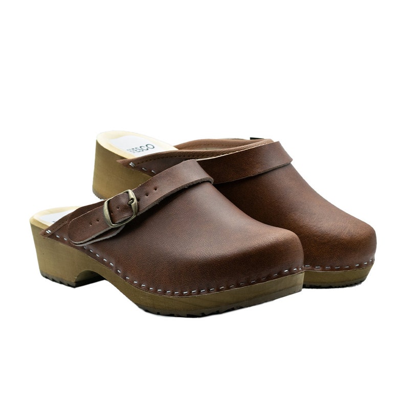 Classic Swedish wooden clogs made of natural cowhide leather in antic brown color and light wooden bottom with orthopedic profile. The upper has a strap with a buckle to adjust the length of the fastening on the heel. Available in women and men size