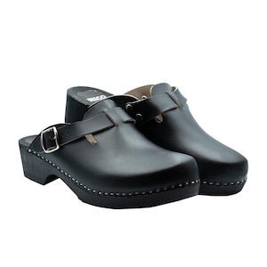 Pode incluir: Um par de tamancos de couro preto com fecho de fivela. Os tamancos têm sola de madeira e são presos com uma fivela prateada. Estes tamancos clássicos são adequados para uso casual.