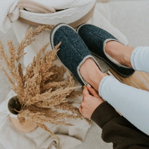 May include: A pair of dark blue fuzzy slippers with a light grey lining. The slippers are being worn by a person with light blue jeans.