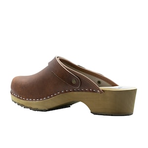 Classic Swedish wooden clogs made of natural cowhide leather in antic brown color and light wooden bottom with orthopedic profile. The upper has a strap with a buckle to adjust the length of the fastening on the heel. Available in women and men size
