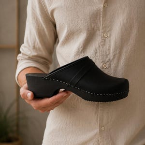 Könnte beinhalten: Schwarze Clogs mit runder Spitze und dicker Sohle. Der Schuh hat einen Riemen über der Oberseite und wird von einer Person gehalten, die ein beiges Hemd mit Knöpfen trägt. Die Clogs haben weiße Nähte an den Rändern.