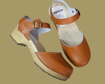 May include: A pair of brown leather clogs with wooden soles and ankle straps. The clogs have a buckle closure and are labeled "Bresco". The wooden soles are approximately 3 inches high.