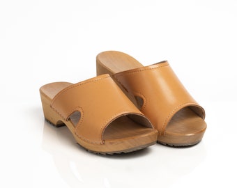 May include: A pair of light brown clogs with open toes and wooden soles. The clogs have a leather upper with a cutout design on the sides. The wooden soles are light brown and have a slight heel. The clogs are set against a white background.