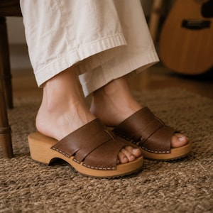 Könnte beinhalten: Ein Paar braune Ledersandalen zum Hineinschlüpfen mit breiten Riemen über dem Spann, offener Zehenpartie und hellen Holzsohlen mit sichtbaren Nähten an den Rändern. Die Sandalen werden zu einer locker sitzenden, hellen Stoffhose getragen.