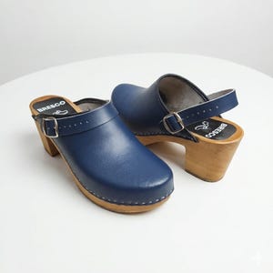 May include: A pair of navy blue clogs with wooden heels and soles. The clogs have a strap around the ankle and a buckle closure. The brand name "Bresco" is visible on the insole. The heels are approximately 3 inches high.