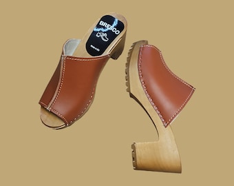 May include: A pair of brown leather clogs with wooden heels and a peep toe. The clogs have a stitched seam around the top edge and the brand name "BRESCO" is printed on the insole. Made in EU.