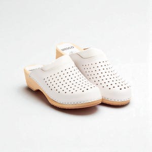 May include: A pair of white leather clogs with a perforated design. The clogs have a wooden sole and a small heel. The brand name "Bresco" is visible on the side of the clogs.