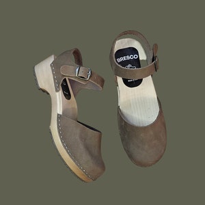 May include: A pair of brown leather clogs with a wooden sole and an ankle strap. The clogs have a brand label that says "BRESCO" on the strap.