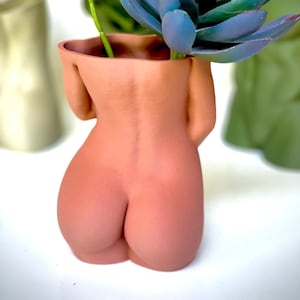 K&ouml;nnte beinhalten: Ein terracottafarbener Keramik-Pflanztopf in Form eines menschlichen Torsos mit einer roten und gr&uuml;nen Blumenanordnung im Inneren.