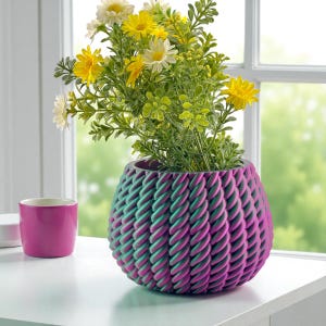 May include: A decorative planter with a rope-like texture, featuring a gradient of teal and magenta. The planter holds a vibrant arrangement of yellow and white flowers with green foliage. A small magenta cup sits nearby.