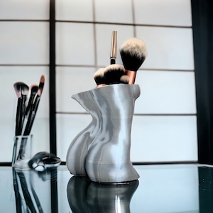 May include: A silver-colored, 3D-printed makeup brush holder shaped like a human torso. The holder has a smooth, textured surface and is filled with makeup brushes.