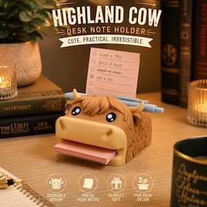 May include: A brown Highland cow desk note holder featuring a light tan muzzle, black and white eyes, and horns. It holds a stack of pink sticky notes in its mouth and a light blue pen across its head. A pink note with the text "PLANT A TREE", "BUILD A HOUSE", "RAISE A CHILD", and "GET A DOG" is inserted into the top.