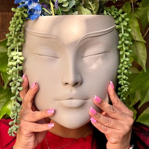 May include: A gray ceramic planter shaped like a human face with closed eyes. The planter is filled with green succulents and blue flowers.