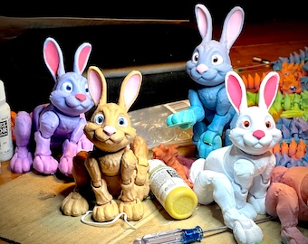 May include: Four articulated bunny rabbit figurines, painted in different colors. The bunnies are posed in various positions, with one sitting, one standing, and two in a more relaxed pose. The bunnies are made of a smooth, plastic-like material.