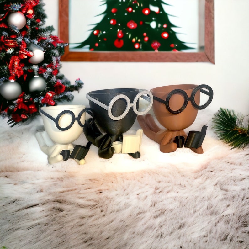 May include: Three ceramic planters shaped like people wearing glasses and reading books. The planters are black, white, and brown.