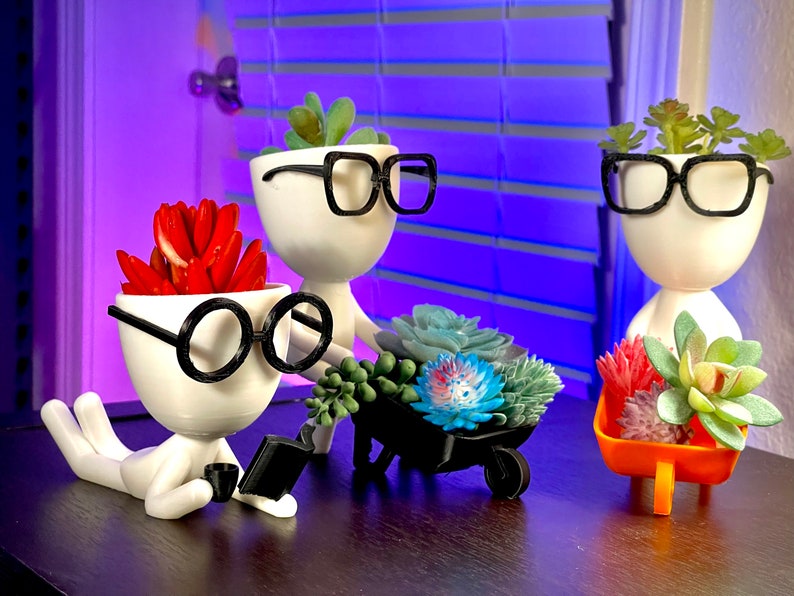 May include: Three white ceramic planters shaped like people with black glasses and different succulents. One planter is holding a small black wheelbarrow filled with succulents.