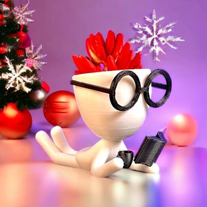 May include: A white planter shaped like a person wearing black glasses, reading a book, with a red poinsettia plant inside.