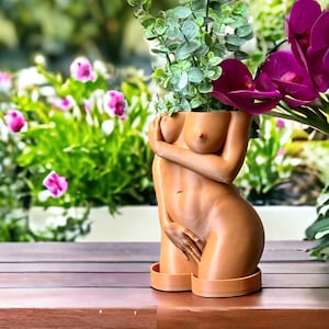 K&ouml;nnte beinhalten: Ein hautfarbener Pflanztopf in Form eines menschlichen Torsos mit einer gr&uuml;nen Pflanze und rosa Bl&uuml;ten, die aus der Oberseite wachsen.
