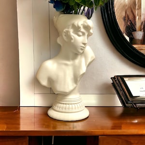 Puede incluir: Jarrón de cerámica blanca en forma de busto de mujer con un rostro esculpido y una base decorativa. El jarrón está lleno de flores azules y verdes.