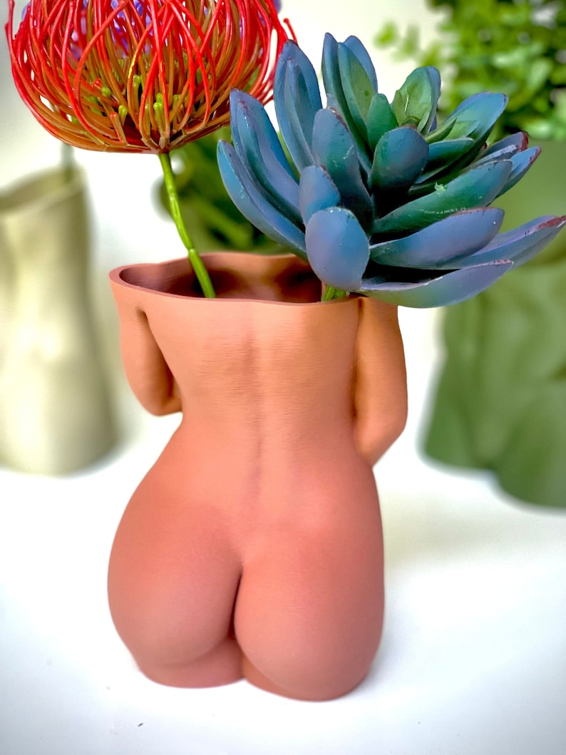 K&ouml;nnte beinhalten: Ein terracottafarbener Keramik-Pflanztopf in Form eines menschlichen Torsos mit einer roten und gr&uuml;nen Blumenanordnung im Inneren.