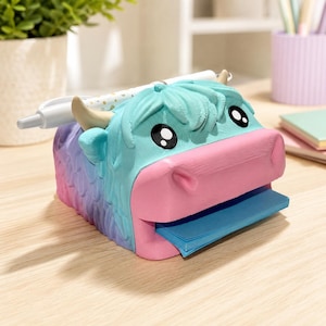 May include: A desk organizer shaped like a highland cow with a gradient blue and pink body. The cow has a light blue head, a pink snout, and two small horns. It features a slot for a pen on top and a dispenser for blue sticky notes in the mouth.