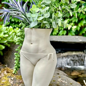 May include: A white ceramic vase shaped like a human torso, with a simple bikini design, holding a bouquet of green and purple artificial plants.