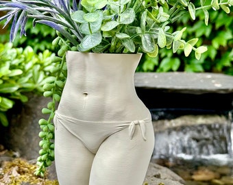 May include: A white ceramic vase shaped like a human torso, with a simple bikini design, holding a bouquet of green and purple artificial plants.