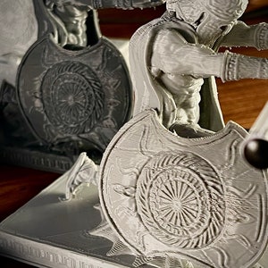 May include: A gray 3D-printed sculpture of a Spartan warrior, complete with helmet, shield, and cape. The shield features a detailed sunburst design. The figure is posed in a dynamic stance, suggesting action. The sculpture is set on a rectangular base.