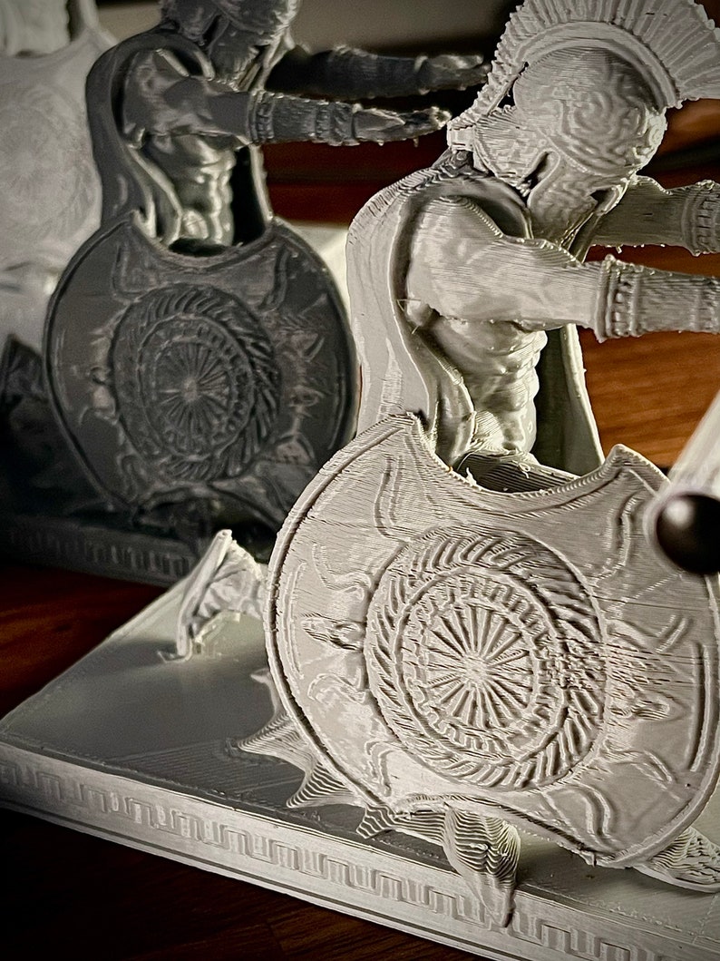 May include: A gray 3D-printed sculpture of a Spartan warrior, complete with helmet, shield, and cape. The shield features a detailed sunburst design. The figure is posed in a dynamic stance, suggesting action. The sculpture is set on a rectangular base.