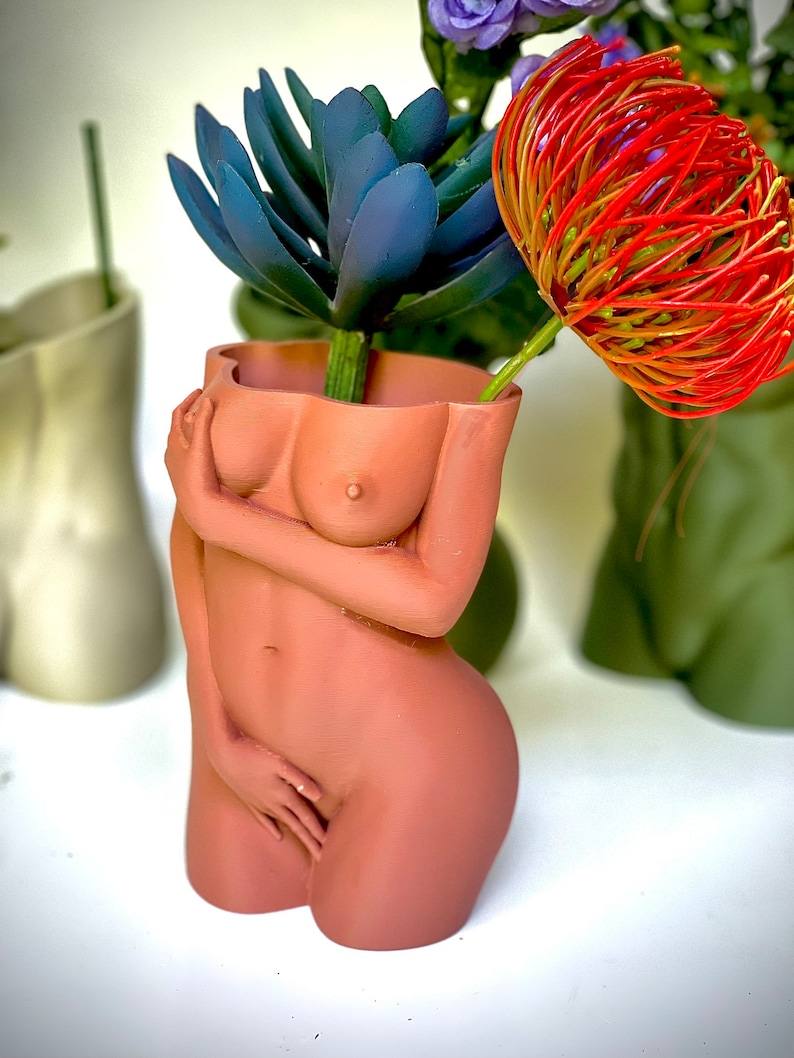 K&ouml;nnte beinhalten: Eine terracottafarbene Keramikvase in Form eines weiblichen Torsos mit einer blauen Sukkulente und einer roten Blume darin.