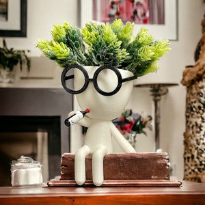 Peut inclure: Un pot en céramique blanche en forme de personne portant des lunettes rondes noires et tenant une petite bouteille rouge et blanche. Le pot a des plantes vertes feuillues qui poussent sur le dessus.