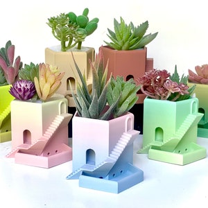 May include: A collection of colorful, miniature architectural planters, each with a unique gradient color scheme. The planters feature a square design with a staircase and archway, and are filled with various faux succulents.