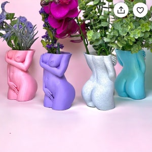K&ouml;nnte beinhalten: Vier Keramik-Blument&ouml;pfe in Form von menschlichen Figuren, jede in einer anderen Farbe: rosa, lila, hellblau und wei&szlig;. Die T&ouml;pfe sind mit gr&uuml;nen Pflanzen und Blumen gef&uuml;llt.