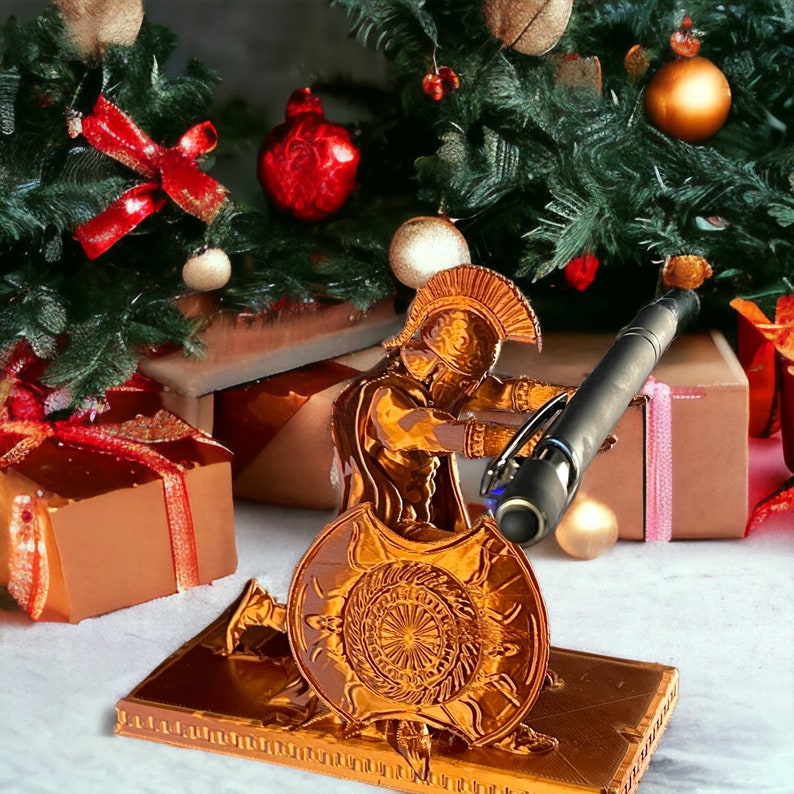 May include: A gold-colored Spartan warrior figurine holding a black pen, with a shield and a rectangular base. The figurine is set against a backdrop of a Christmas tree, wrapped gifts, and ornaments.