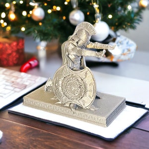 May include: A silver-colored figurine of a Spartan warrior with a shield and helmet. The warrior is kneeling on a rectangular base. The background includes a Christmas tree with ornaments and lights.