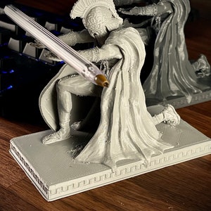 May include: A gray 3D-printed figurine of a kneeling warrior in classical armor, holding a pen. The figurine is on a rectangular base with a Greek key border. Another similar figurine is in the background.