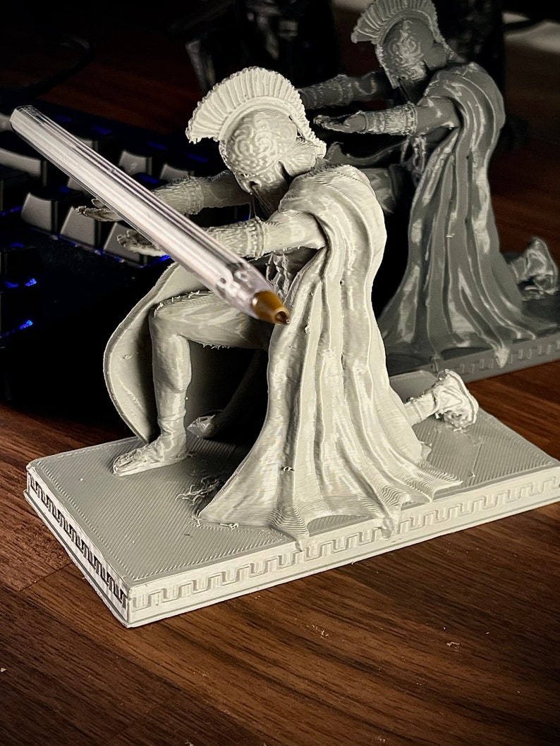 May include: A gray 3D-printed figurine of a kneeling warrior in classical armor, holding a pen. The figurine is on a rectangular base with a Greek key border. Another similar figurine is in the background.