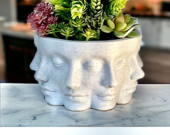 May include: White ceramic planter with four faces, each with closed eyes and a slightly parted mouth. The planter is filled with green succulents and a small cluster of pink flowers.