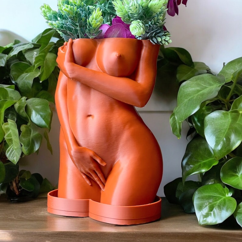 K&ouml;nnte beinhalten: Ein terracottafarbener Pflanztopf in Form eines menschlichen Torsos, aus dem oben eine gr&uuml;ne, belaubte Pflanze w&auml;chst. Der Topf hat eine glatte, matte Oberfl&auml;che.