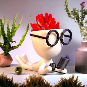May include: A white ceramic planter shaped like a person wearing glasses, holding a book and a cup. The planter has red flowers growing out of the top.
