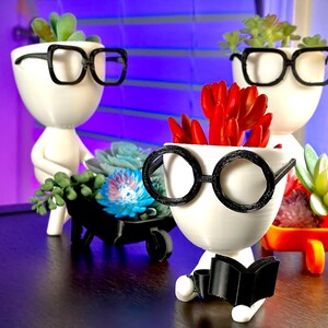 May include: Three white ceramic planters shaped like people with black-rimmed glasses. The planters are holding succulents and are sitting on a dark wood surface. One planter is holding a book.