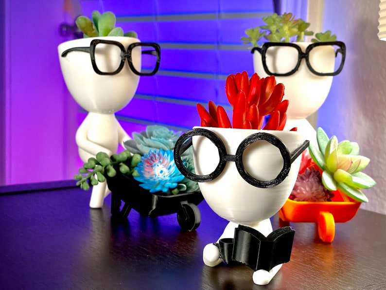 May include: Three white ceramic planters shaped like people with black-rimmed glasses. The planters are holding succulents and are sitting on a dark wood surface. One planter is holding a book.