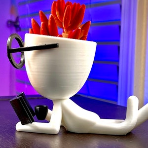 May include: White 3D printed planter shaped like a person wearing glasses, holding a book and a cup. The planter has a red succulent plant inside.
