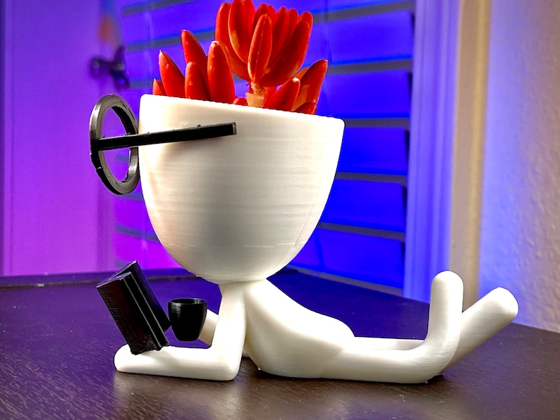 May include: White 3D printed planter shaped like a person wearing glasses, holding a book and a cup. The planter has a red succulent plant inside.