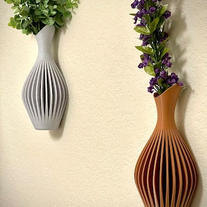 May include: Two decorative wall vases with a unique, geometric design. The vase on the left is light grey and holds a sprig of green leaves. The vase on the right is brown and holds a sprig of purple flowers.
