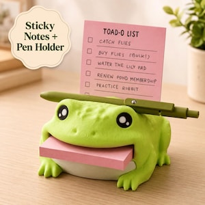 May include: A light green frog-shaped desk organizer featuring a slot for a stack of pink sticky notes in its mouth and a horizontal olive green pen holder across its back. A pink notepad stands upright with the text TOAD-O LIST and tasks.